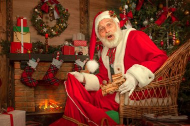 Noel Baba hediye dolu bir çantayla şömine ve Noel ağacının yanında. Ahşap evin içindeki şenlikli iç mekan, Noel 'in neşeli ruhu. Gerçek beyaz sakallı son sınıf öğrencisi Noel Baba kostümü giyiyor..