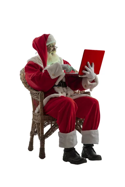 サンタクロースの白い背景に孤立した シニア男性俳優古いです男とともに本当の白いひげでザ父クリスマス座っていますでザウィッカー柳の椅子とともに赤ラップトップ — ストック写真