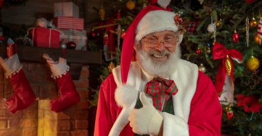 Noel Baba şöminenin ve Noel ağacının yanında el hareketi yapıyor. Ahşap evin içindeki şenlikli iç mekan, Noel 'in neşeli ruhu. Gerçek beyaz sakallı son sınıf öğrencisi Noel Baba kostümü giyiyor..