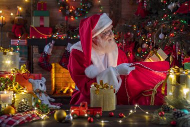 Noel Baba hediye dolu bir çantayla şömine ve Noel ağacının yanında. Ahşap evin içindeki şenlikli iç mekan, Noel 'in neşeli ruhu. Gerçek beyaz sakallı son sınıf öğrencisi Noel Baba kostümü giyiyor..