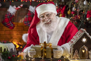 Noel Baba hediye kutusuyla şömine ve Noel ağacının yanında duruyor. Ahşap evin içindeki şenlikli iç mekan, Noel 'in neşeli ruhu. Gerçek beyaz sakallı son sınıf öğrencisi Noel Baba kostümü giyiyor..