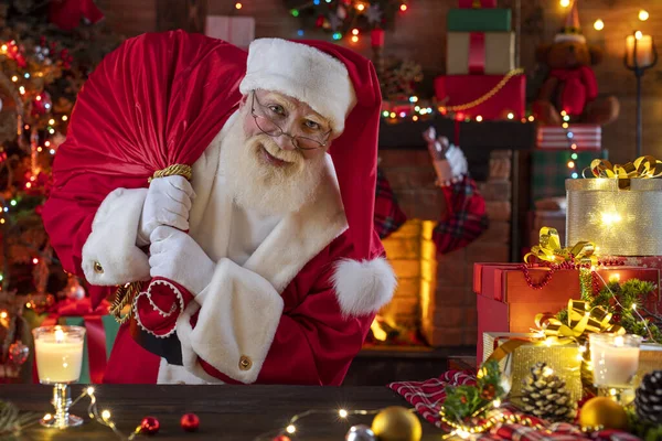 Noel Baba hediye dolu bir çantayla şömine ve Noel ağacının yanında. Ahşap evin içindeki şenlikli iç mekan, Noel 'in neşeli ruhu. Gerçek beyaz sakallı son sınıf öğrencisi Noel Baba kostümü giyiyor..