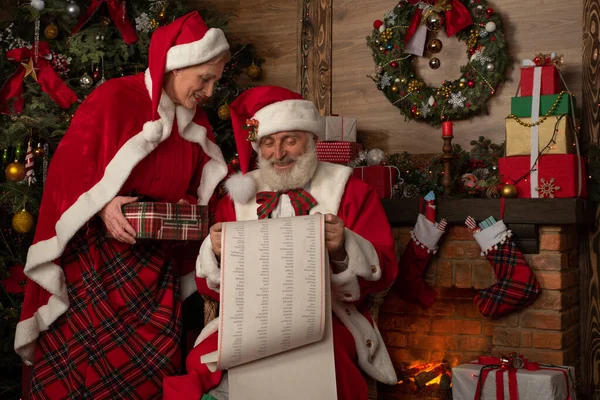 Noel Baba ve karısı Noel Anne şöminenin ve Noel ağacının yanında. Ahşap evin içinde şenlikli bir iç mekan, yeni yılın Noel Ruhu. Gerçek beyaz sakallı son sınıf öğrencisi Noel Baba kostümü giyiyor.