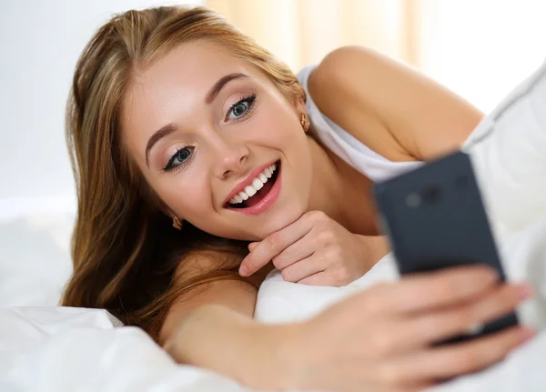 젊은 아름 다운 금발의 여 자가 핸드폰을 들고 침대에 누워 — 스톡 사진