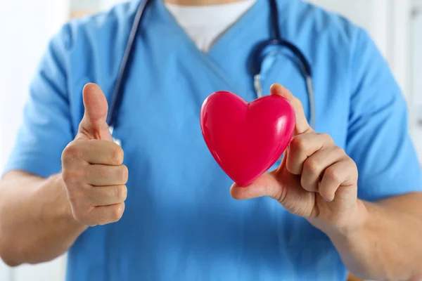 Чоловік-лікар у блакитній формі тримає червоне іграшкове серце і показує добре — стокове фото