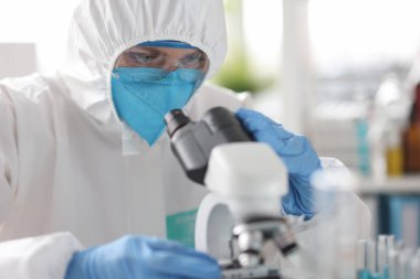 Koruyucu giysi ve solunum cihazında çalışan bir bilim adamı laboratuvarda mikroskoptan bakıyor.