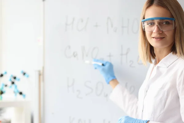 Mujer científica en guantes protectores mostrando fórmulas químicas en pizarra — Foto de Stock