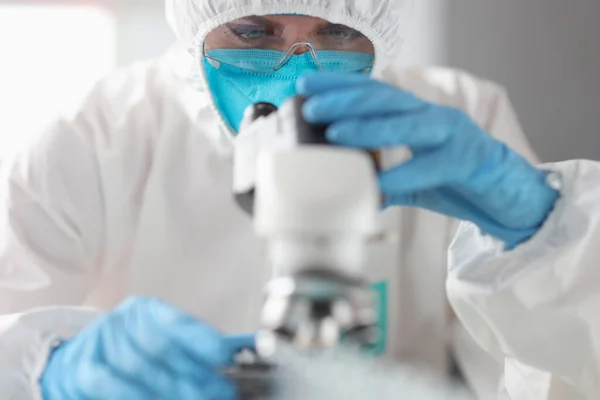 Koruyucu kıyafetli ve gözlüklü doktor mikroskopla bakıyor.