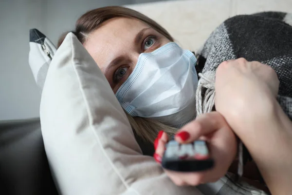 保護医療マスクの病気の女性はソファに横たわっており、リモートコントロールを保持しています — ストック写真