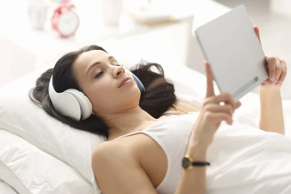 Woman with headphones lying on bed and holding tablet — Stock Photo, Image