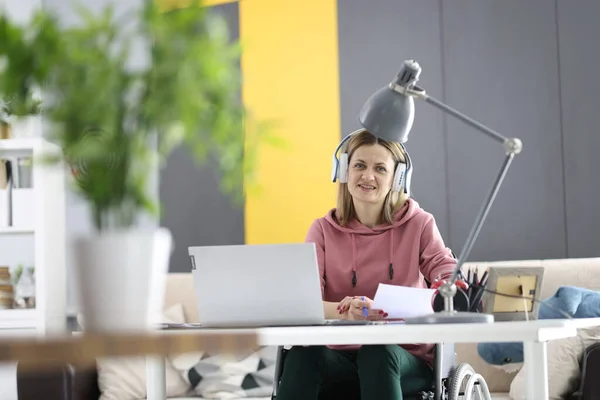 Žena na vozíčku sedí u pracovního stolu a nosí sluchátka. — Stock fotografie