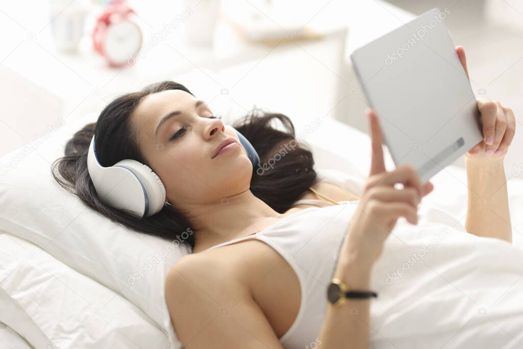 Woman with headphones lying on bed and holding tablet