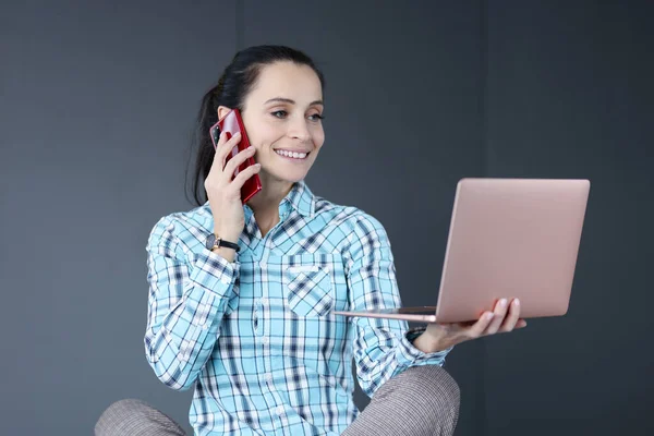 Mujer joven sosteniendo el ordenador portátil en sus manos y hablando por teléfono —  Fotos de Stock