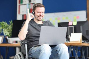 Kulaklıklı ve tekerlekli sandalyedeki mikrofonlu adam dizüstü bilgisayara iyi bir hareket gösteriyor.