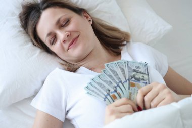 Elinde dolarlar olan beyaz yatakta uyuyan bir kadın.