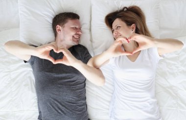 Kadın ve erkek yatakta yatar ve elleriyle kalplerini gösterir.