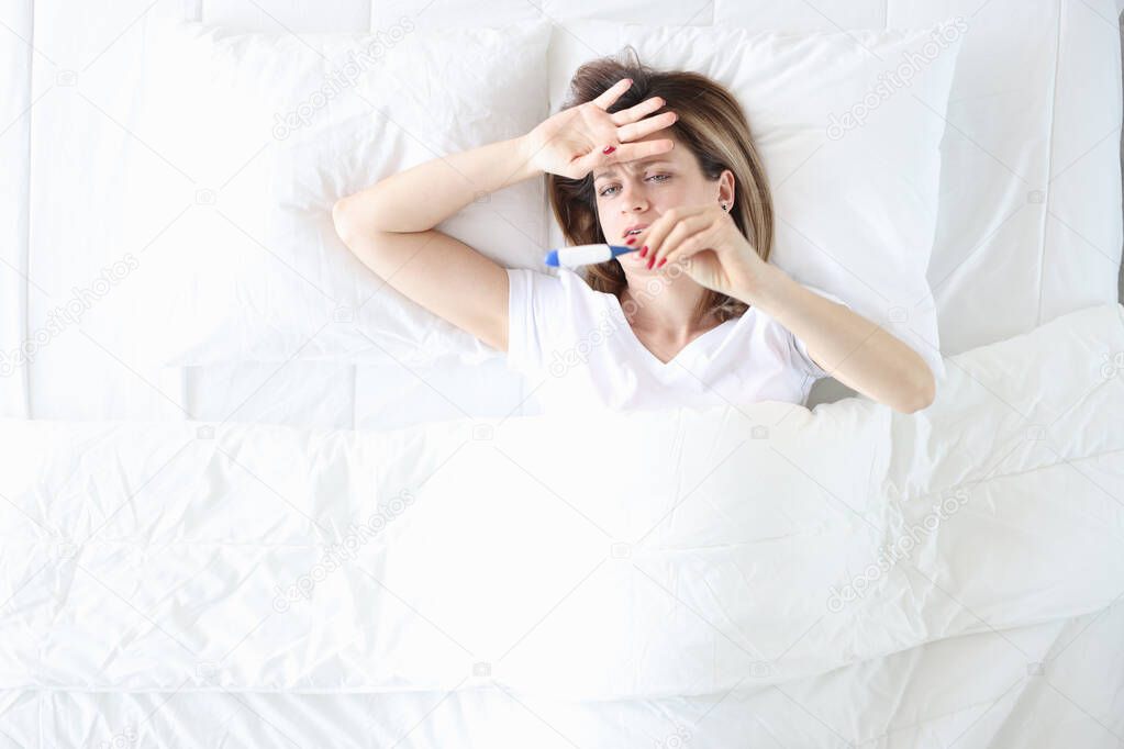 Sick woman lies in bed with thermometer closeup