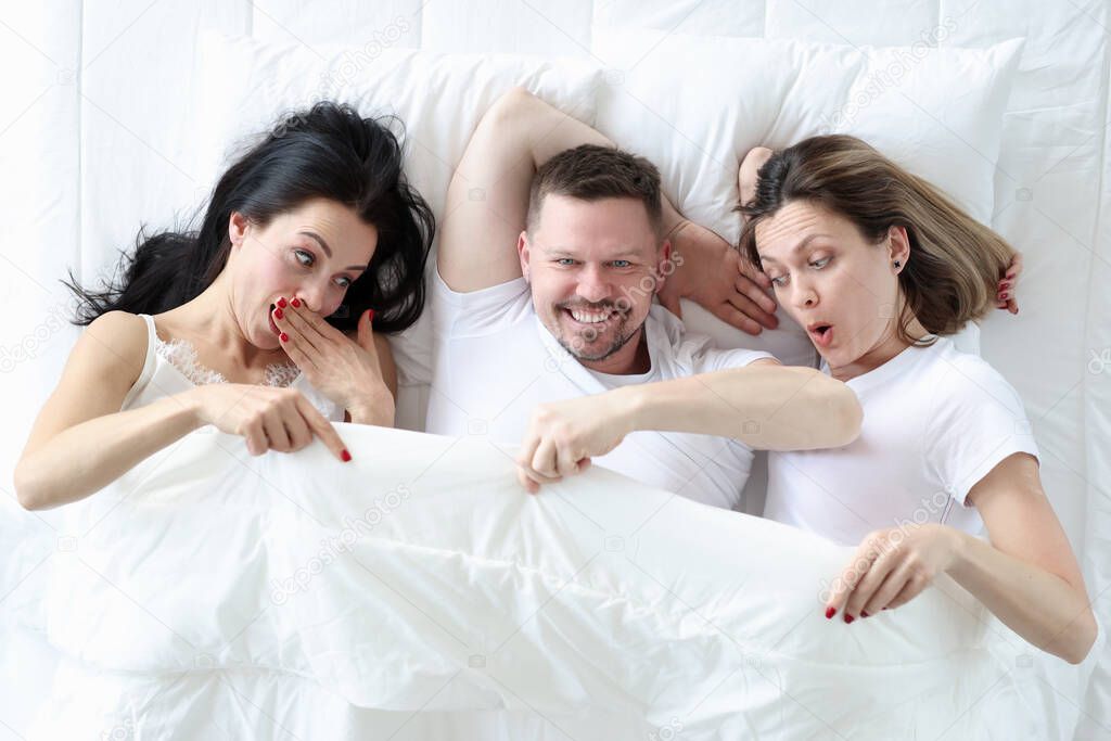 Man lying with two lovers in bed. Women looking under cover and wondering