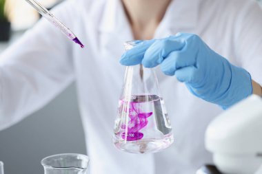 Scientist in laboratory holds transparent flask of liquid and digs purple reagent into it