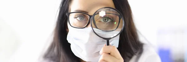 Doctor in protective medical mask looks through magnifying glass — Stock Photo, Image