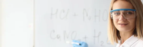 Женщина-ученый в защитных перчатках показывает химические формулы на доске — стоковое фото