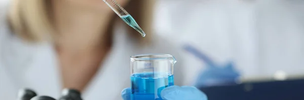 Wissenschaftlerin tropft blaue Flüssigkeit aus Pipette in Kolben im Labor — Stockfoto