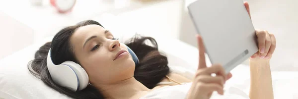 Wanita dengan headphone berbaring di tempat tidur dan memegang tablet — Stok Foto