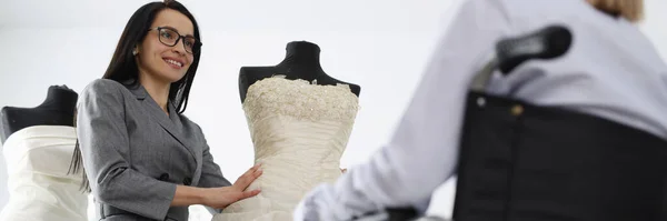 Woman consultant demonstrates wedding dress on mannequin to woman in wheelchair — Stock Photo, Image