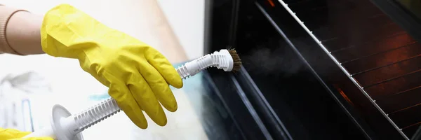 Female hands in yellow gloves are holding a steam cleaner and cleaning oven