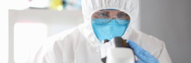 Koruyucu giysili, maskeli ve gözlüklü doktor mikroskopla bakıyor.