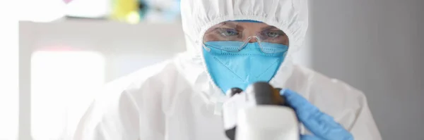 Doctor in protective medical suit, mask and glasses looks through microscope