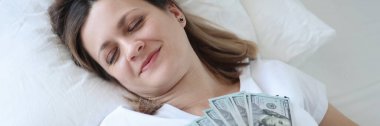 Elinde dolarlar olan beyaz yatakta uyuyan bir kadın.