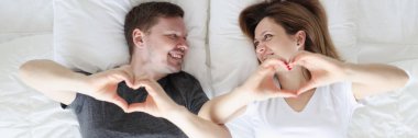 Kadın ve erkek yatakta yatar ve elleriyle kalplerini gösterir.