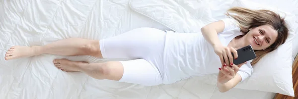 Mulher de pijama branco deitada na cama e tendo vista superior selfie — Fotografia de Stock