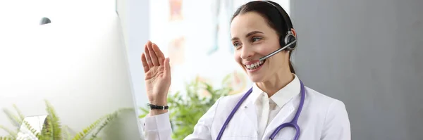 Doctor with headphones and microphone waving hand at computer screen — стоковое фото