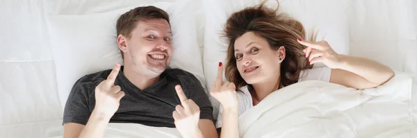 Smiling man and woman lie in bed and call fak gesture — Stock Photo, Image