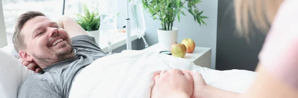 Sorrindo homem encontra-se na cama do hospital sua mão é realizada pela mulher — Fotografia de Stock