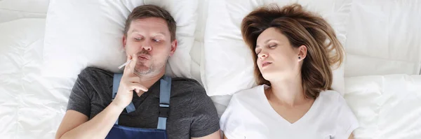 Smoking man with cigarette lies in bed with his sleeping wife — Stock Photo, Image