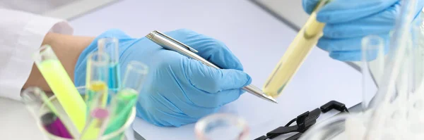 Químico cientista segurando tubo de teste com líquido amarelo em suas mãos e escrevendo em documentos close-up — Fotografia de Stock