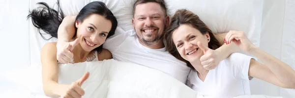 Dos mujeres sonrientes con hombre yacen en la cama y levantan los pulgares —  Fotos de Stock