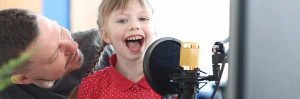 En liten flicka som sitter i sina fäders knä och sjunger sång i mikrofon — Stockfoto