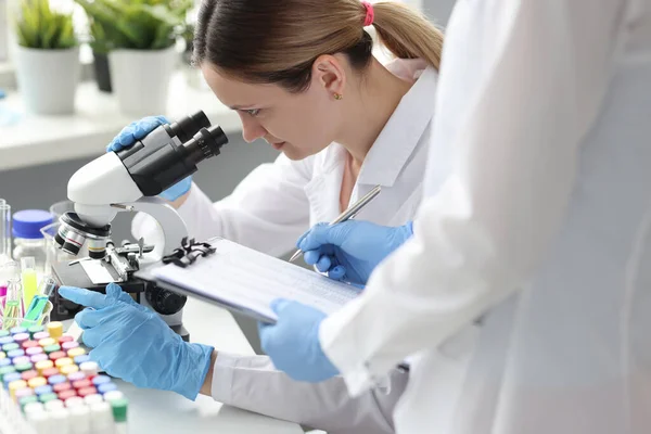 Mujer científica realiza prueba de composición química de la sustancia bajo el microscopio asistente escribe datos — Foto de Stock