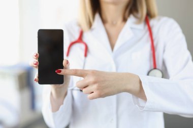 Beyaz önlüklü doktor akıllı telefonu tutuyor ve ona yakın çekimde işaret ediyor.
