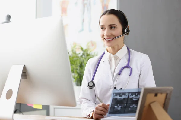 Arzt mit Kopfhörer und Mikrofon hält Röntgenbild vor Computerbildschirm — Stockfoto