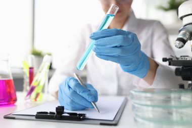 Kadın bilim adamı eldiven içinde mavi sıvıyla test tüpü tutuyor ve elde edilen kimyasal verileri yazıyor.