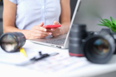 Kadın akıllı telefonu elinde tutarken kameradan alınan lensin yanında dizüstü bilgisayar duruyor.