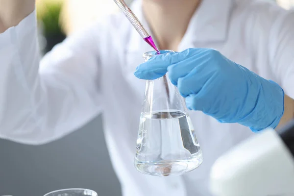 Naukowiec w rękawiczce trzyma kolbę wody i wrzuca do niej fioletowy płyn z pipety. — Zdjęcie stockowe