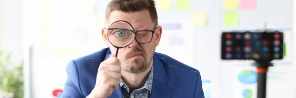 Young business coach holding magnifying glass near eye in front of mobile phone camera — Stock Photo, Image