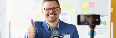Smiling businessman holds thumbs up and records business training on video