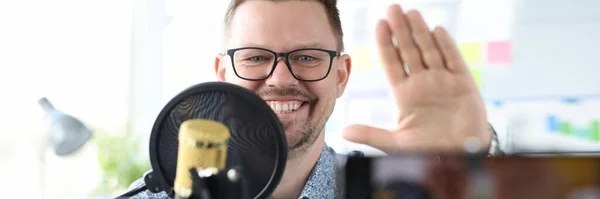 Smiling man sits in front of a microphone and waves at camera with his hand — Stock Photo, Image